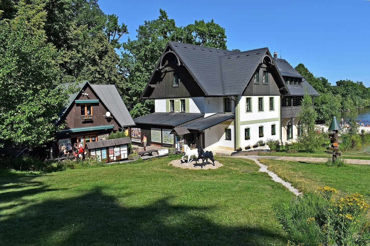 Autokamp Sedmihorky  - Blick auf zwei in naturfarben gehaltene Gebäude des Campingplatzes, davor eine grüne Wiese und im Hintergrund Strand und See