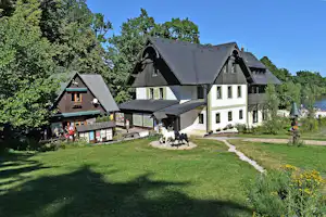 Autokamp Sedmihorky - Blick auf zwei in naturfarben gehaltene Gebäude des Campingplatzes, davor eine grüne Wiese und im Hintergrund Strand und See
