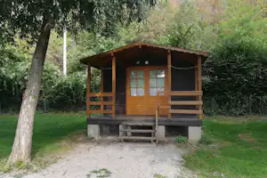 Holzhütte auf einem Campingplatz im Grünen, mit Veranda, Holzgeländer und kleinem Eingang, umgeben von Bäumen und Rasen.