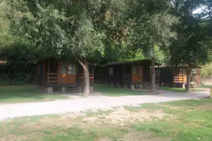 Holzbungalows auf einem Campingplatz im Grünen, mit Veranda, Sitzbank und großen Bäumen im Hintergrund.