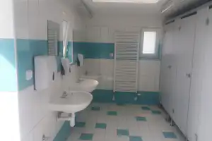 Gemeinschaftsbadezimmer mit mehreren Waschbecken, weißen Fliesen und türkisfarbenen Wandstreifen, daneben Duschbereich mit Glastür.