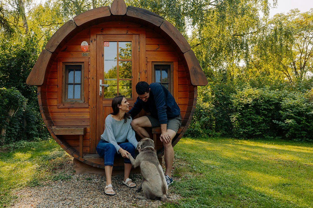 Runde Holzblockhütte im Garten, davor sitzen eine Familie und ein Hund auf dem Kiesweg.