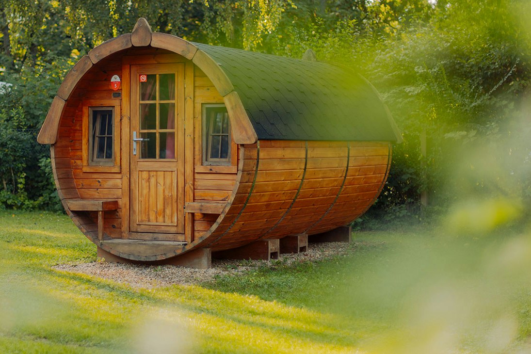 Holzfass-Sauna auf einem Campingplatz im Grünen, mit Eingangstür und Fenstern, umgeben von Rasen und Bäumen.