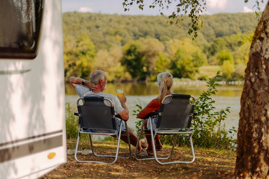 Zwei Personen sitzen auf Campingstühlen am Wohnwagen, im Hintergrund ein See und bewaldete Hügel.