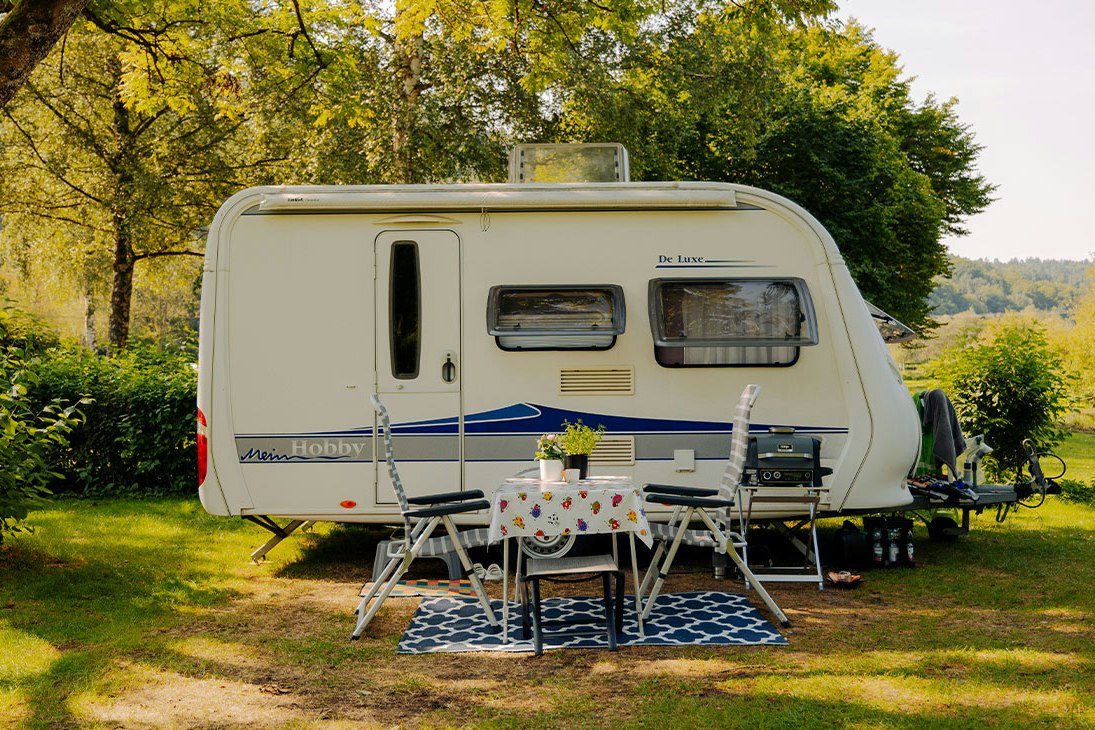 Wohnwagen auf einem Campingplatz-Stellplatz im Grünen, mit Vorzelt, Tisch und Stühlen vor dem Fahrzeug, umgeben von Bäumen.