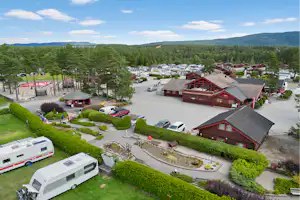 Campinganlage mit roten Holzhaeusern, Stellplaetzen und Wohnmobilen, umgeben von Baeumen und gruenen Hecken.