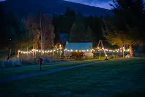Campingplatz am Abend mit beleuchteten Lichterketten, Zelt und Holzzaun im Hintergrund, Bäume und Hügel im Dunkeln.