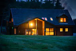 Holzhaus auf dem Campingplatz am Abend, beleuchtete Fenster und Eingang, Satteldach und Bäume im Hintergrund.