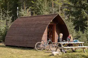 Holzhütte im Campingbereich mit Veranda, Tisch und Fahrrädern davor, umgeben von Bäumen im Garten.