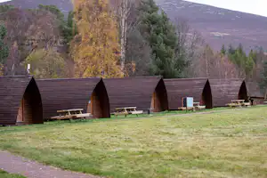 Reihe von Holz-Hütten am Campingplatz mit Sitzbänken und Rasenfläche, umgeben von Bäumen und Hügeln im Hintergrund.