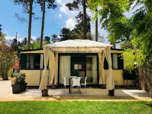 Campingzelt mit Vorhangdach und Terrasse, zwei Stühle und Tisch vor dem Zelt, umgeben von Bäumen und Rasen.