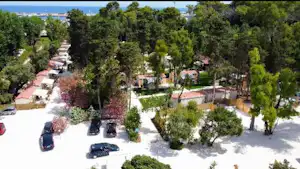 Luftaufnahme eines Campingplatzes mit Stellplätzen, kleinen Häusern und Bäumen, dazwischen gepflasterte Wege und geparkte Autos.