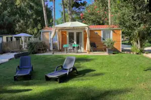 Stellplatz mit Mobilheim und Terrasse im Grünen, zwei Liegestühle auf dem Rasen, Hecken und Bäume im Hintergrund.