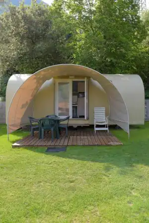Zelt mit Terrasse und Sitzgruppe auf einem Campingplatz, im Hintergrund ein Wohnwagen und Bäume im Grünen.