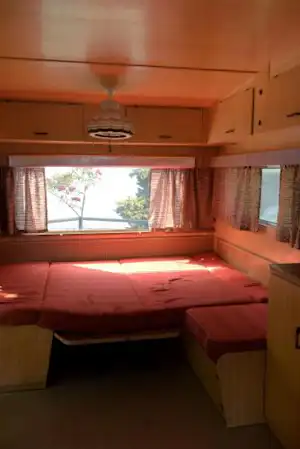 Innenansicht eines Wohnwagens mit Doppelbett, Schrankwand und großen Fenstern, Vorhängen und Blick ins Freie.