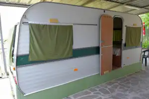 Wohnwagen mit Vorzelt und grünen Stoffbahnen, seitlicher Eingang und Holzverkleidung, gepflasterter Stellplatz am Campingplatz