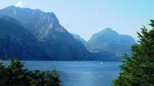 Blick auf einen See mit Segelboot, umgeben von Bergen und grünen Bäumen im Vordergrund.