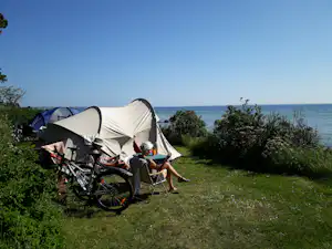 Campingzelt am Meer, davor Fahrräder und Campingstühle auf einer grünen Wiese unter klarem Himmel.