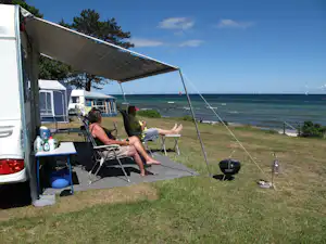 Wohnmobil-Stellplatz am Meer: Markise, Campingstühle und Tisch unter Sonnenschirm, im Hintergrund Zelte und Segelboote.
