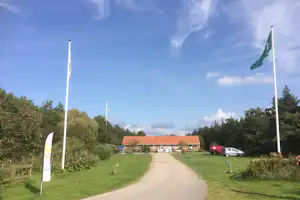 Campingplatzweg mit Rasen und Fahnen, im Hintergrund Gebäude mit roten Dächern und geparkter weißer Wagen.