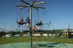 Seilnetz-Spielgerät auf dem Campingplatz, Kinder auf dem Klettergerüst mit bunten Seilen und Netzen vor blauem Himmel.