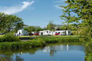 Wohnmobile und Campingbusse auf dem Stellplatz am Fluss, umgeben von Rasen, Bäumen und einem Weg.