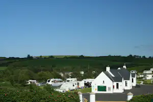 Campingplatz mit Wohnmobilen und Wohnwagen auf Stellplätzen, weiße Gebäude und Dächer im Vordergrund, grüne Hügel und Himmel.