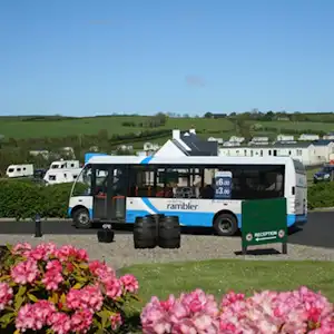 Wohnmobil und Bus an der Rezeption auf einem Campingplatz, im Hintergrund Gebäude und Wohnwagen, davor Blumenbeete.