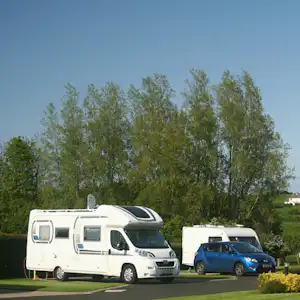 Wohnmobil und blauer Pkw auf einem Campingplatz-Stellplatz, im Hintergrund Bäume und ein weiteres Fahrzeug.