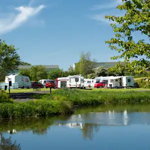 Reihe von Wohnmobilen und roten Autos auf dem Campingplatz am Ufer eines kleinen Gewässers, mit Bäumen im Vordergrund.