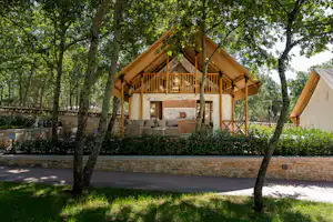 Holz-Chalet mit Veranda und Geländer, umgeben von Bäumen und Hecken, im Campingbereich mit gepflastertem Weg.
