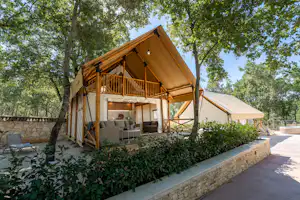 Glamping-Zelt mit Holzveranda, davor Sitzbereich und Grill, umgeben von Hecken und Bäumen am Campingplatz.