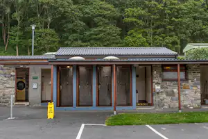 Überdachter Eingang eines Campinggebäudes mit Steinmauer, Holzverkleidung und mehreren Türen, davor Parkplatz und Rasenstreifen.