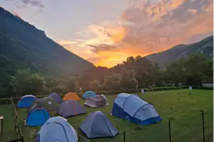Zelte auf einer grünen Wiese im Campingplatz, umgeben von Bergen und Bäumen, mit goldenem Sonnenuntergang am Himmel.