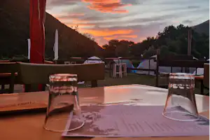 Blick auf Campingzelte auf einer Wiese, umgeben von Bergen und Bäumen, mit Tisch und Gläsern im Vordergrund.