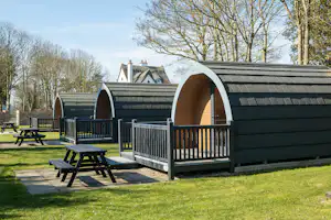 Holzhütten mit Veranda und Sitzgarnitur auf einem gepflegten Campingplatz, umgeben von Bäumen und Wegen.