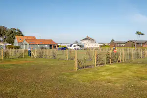 Grüner Campingplatz mit gepflegtem Rasen, Zaun und jungen Pflanzen vor mehreren Gebäuden mit roten Ziegeldächern.