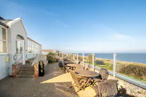 Terrasse am Campingplatz mit Holztisch und Stühlen, Ziegelweg, weißen Gebäuden und Meerblick am Horizont.