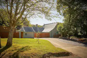 Campinggebäude mit Schieferdach, Kiesweg und Rasenfläche, umgeben von Bäumen und Holzbank am Rand.