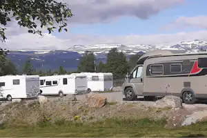 Wohnmobile auf einem Campingplatz, im Hintergrund Berge und Bäume, davor ein Kiesstreifen und Rasen.