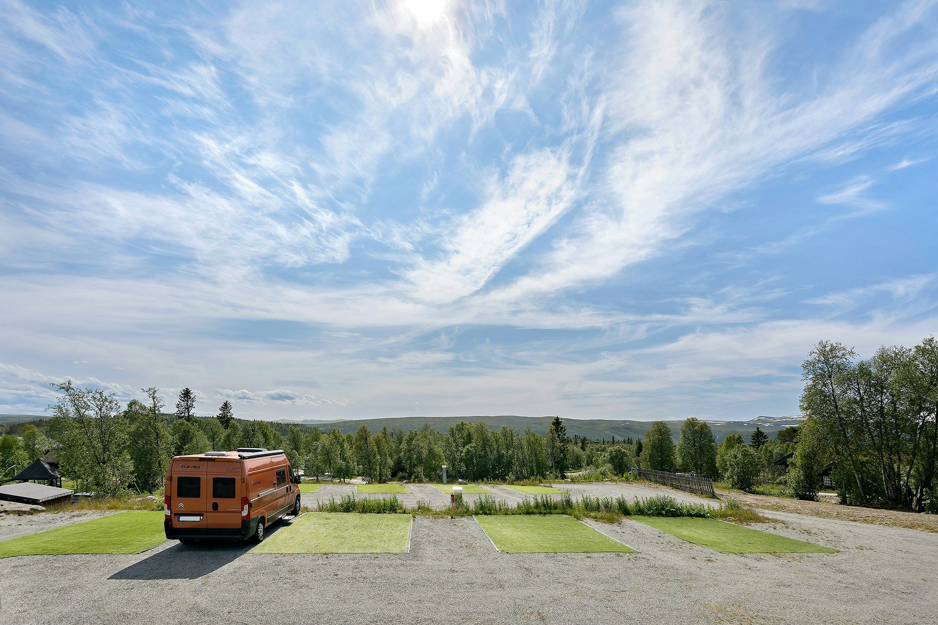 Wohnmobil auf einem Campingplatz mit Rasenflächen und Wegen, im Hintergrund Bäume, Felder und eine weite Landschaft.