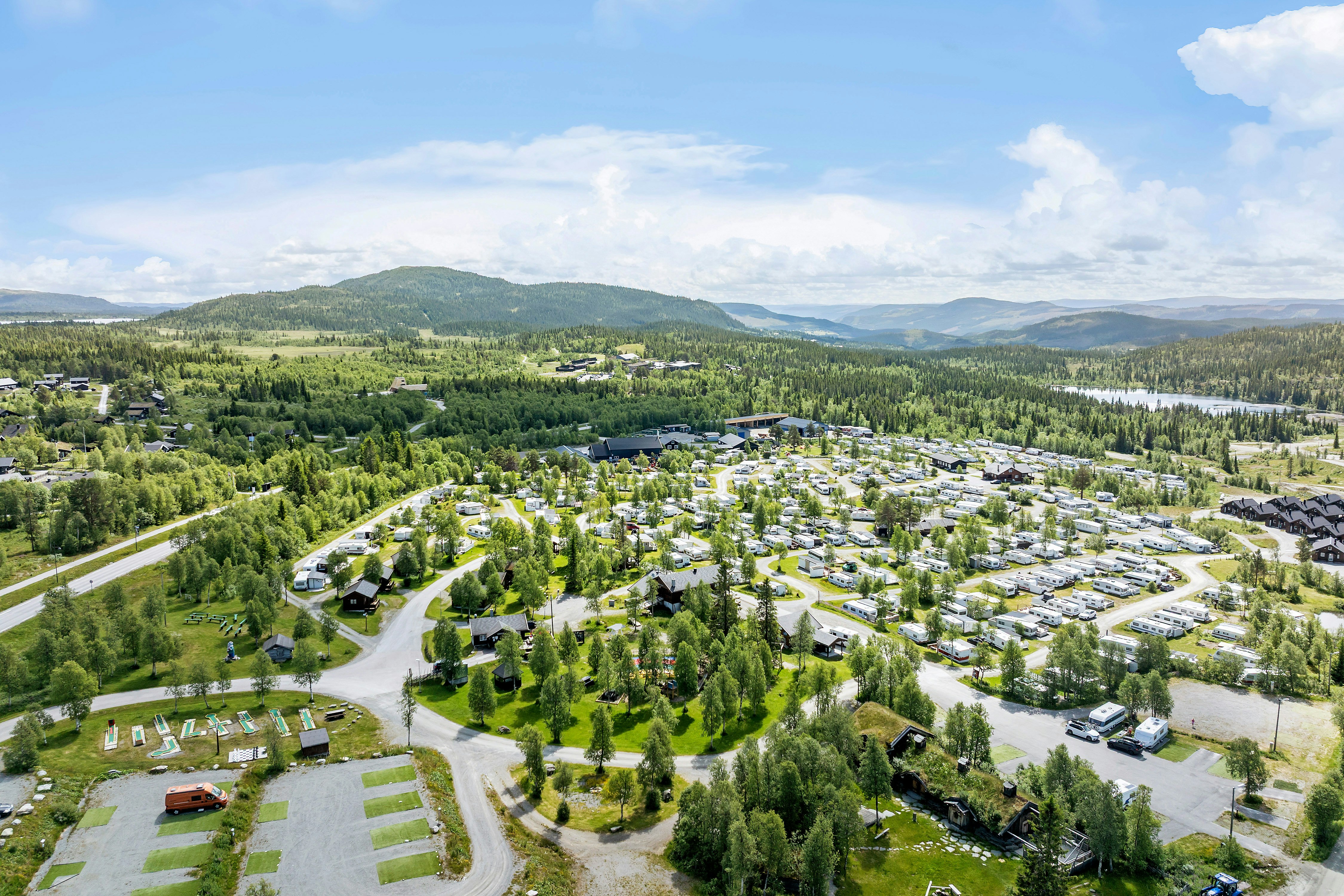 Luftaufnahme eines Campingplatzes mit vielen Stellplätzen, Wohnwagen und Zelten, umgeben von Wäldern und Hügeln.