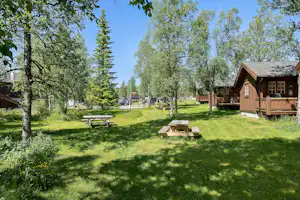 Holzhütten und ein Picknicktisch auf einer grünen Wiese im Campingbereich, umgeben von Bäumen und einem Weg.