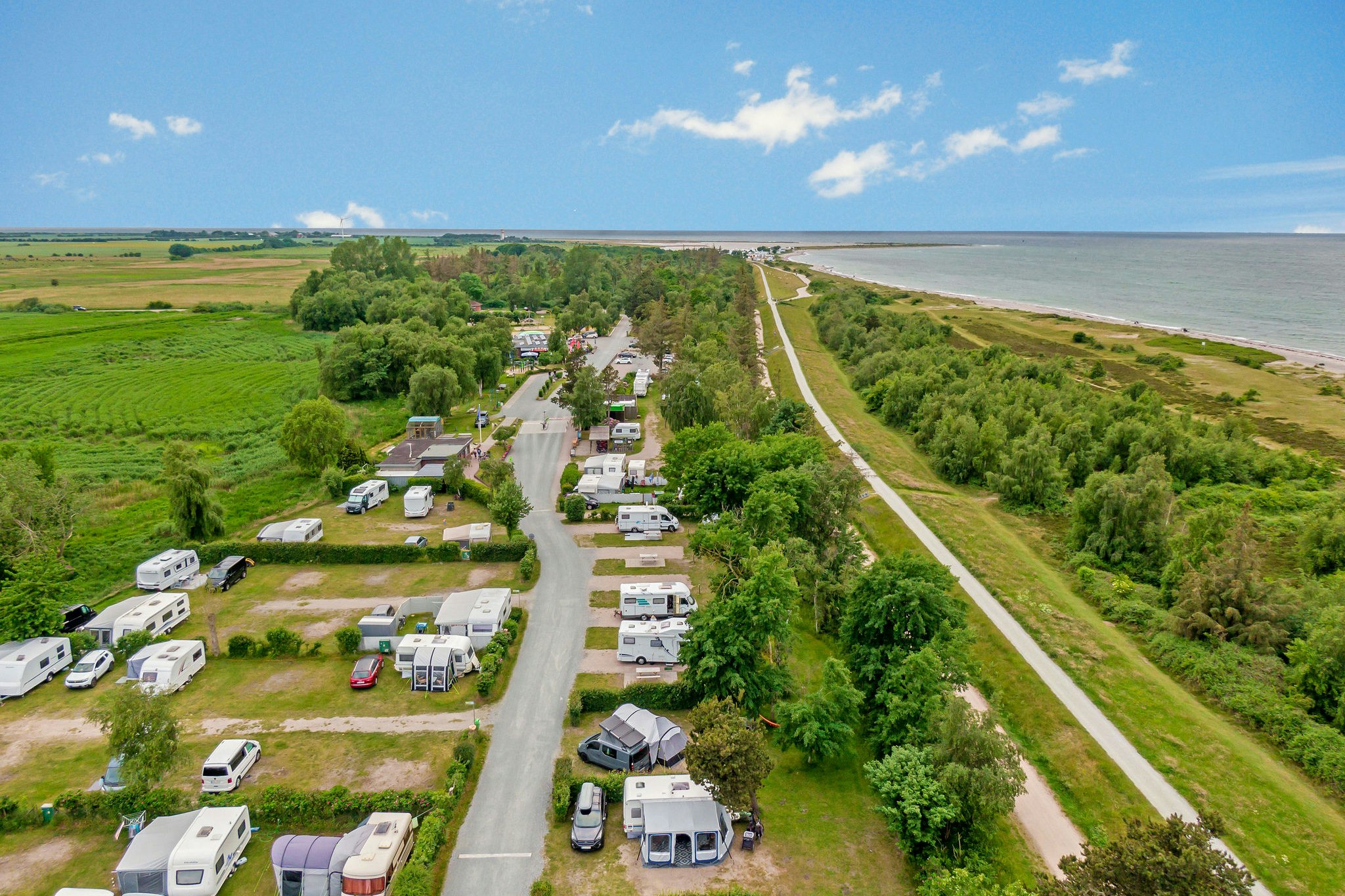 Campingplatz mit Wohnmobilen und Stellplätzen, Feldweg zum Meer, grüne Landschaft und Bäume am Ufer.