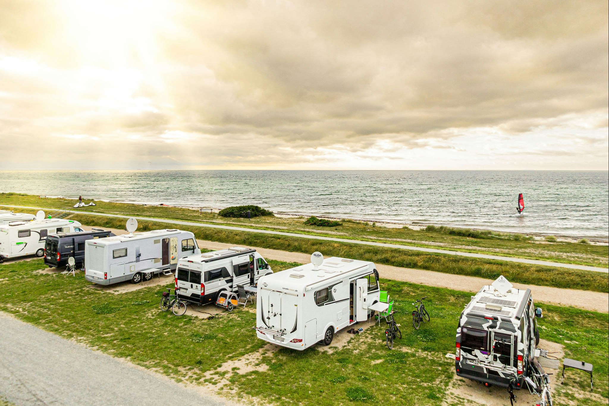 Wohnmobile und Campingfahrzeuge auf einem Stellplatz am Strand, mit Grasfläche, Weg und Blick auf das Meer.