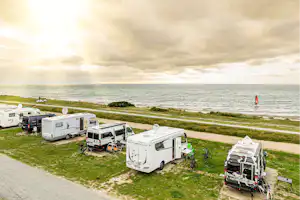 Wohnmobile und Campingfahrzeuge auf einem Stellplatz am Strand, mit Grasfläche, Weg und Blick auf das Meer.