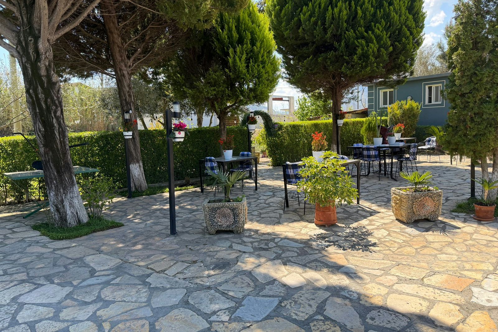 Camping-Terrasse mit Steinterrasse, mehreren Tischen und Stühlen, Töpfen mit Pflanzen und Bäumen neben einem Wohnwagen.