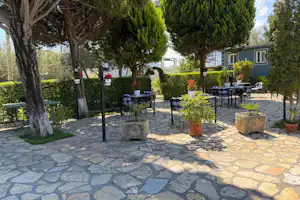 Camping-Terrasse mit Steinterrasse, mehreren Tischen und Stühlen, Töpfen mit Pflanzen und Bäumen neben einem Wohnwagen.