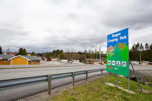 Camping Park Schild am Straßenrand neben einem Metallgeländer, im Hintergrund Wohnhäuser und Bäume entlang der Zufahrt.