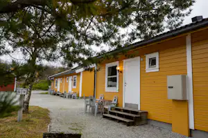 Gelbe Ferienhaus-Reihe mit Terrasse, Sitzbank und Tisch vor den Eingängen, Kiesweg und Bäume im Hintergrund.